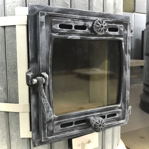 Detail view of the heavy-duty cast iron firebox door with a distressed antique silver finish, mounted on a grey stone Siberia heater.