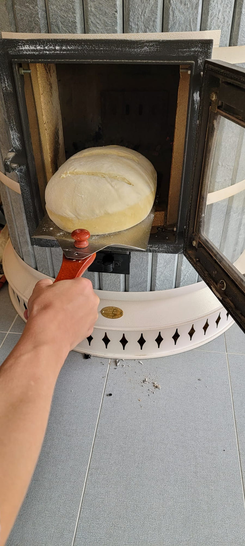 A person using a pizza peel to load a loaf of artisan bread into the hot firebox of a Siberia masonry heater for baking.