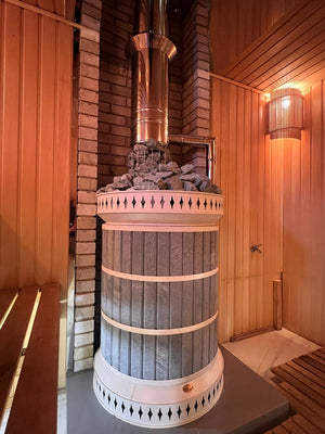 Volcano Blue masonry sauna heater installed in a corner of a sauna room with cream bands and a wire mesh rock basket on the chimney
