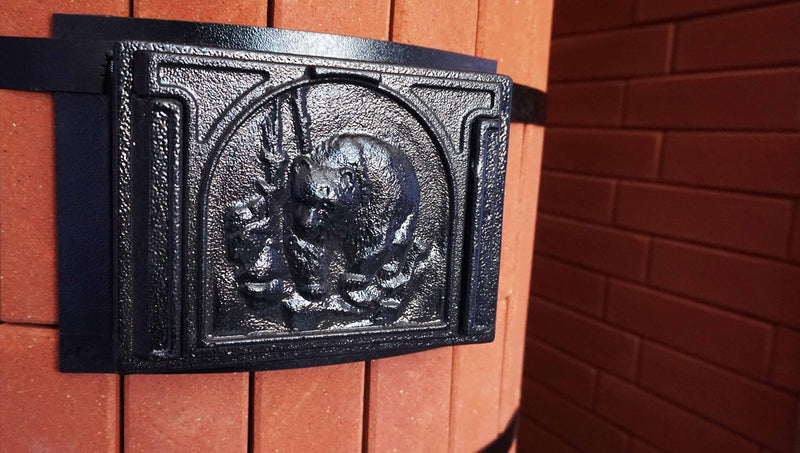 Close-up of a heavy-duty cast iron cleanout door featuring a decorative bear relief sculpture, mounted on a red brick Tundra masonry heater.