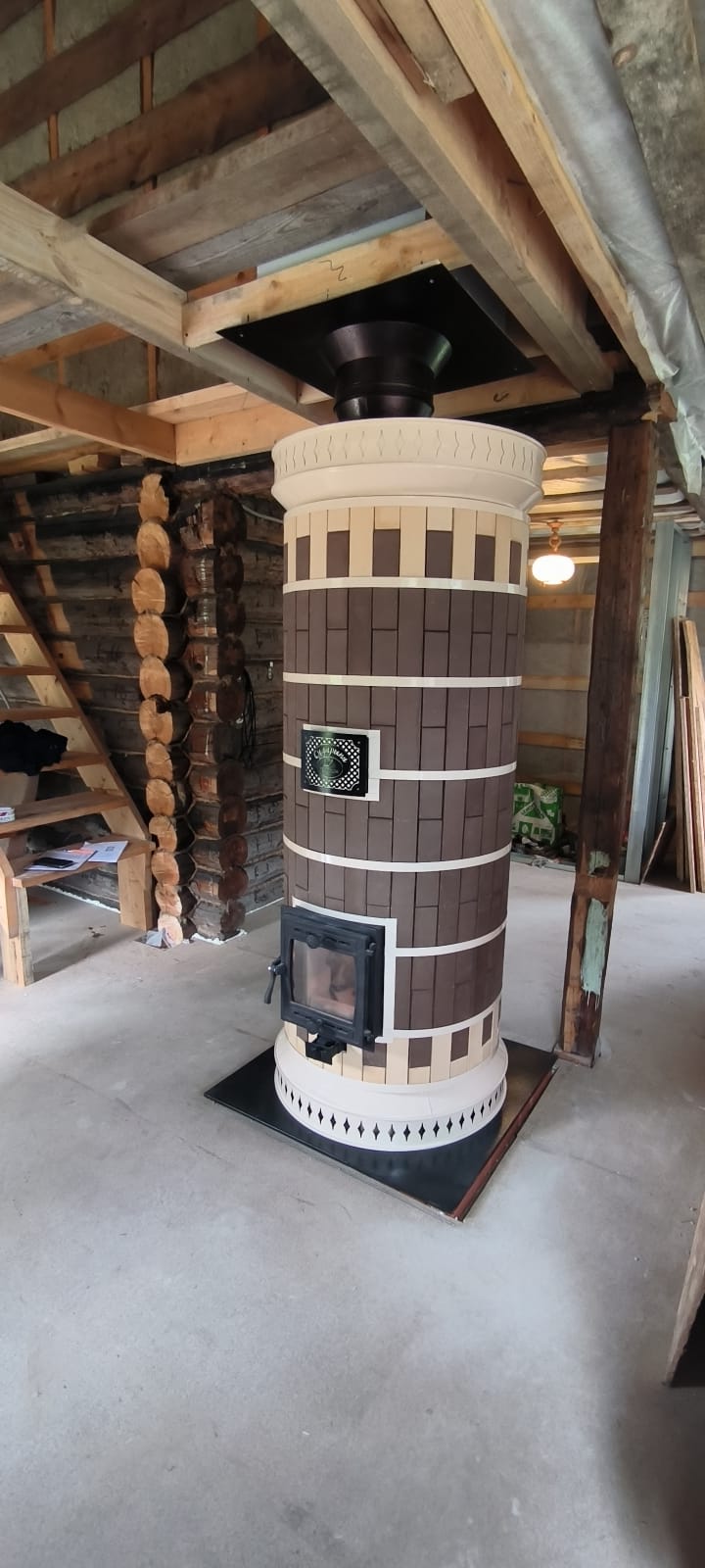 Installation of a DIY Brown Siberia masonry heater kit in a log cabin under construction, showing the heater positioned on a black floor protection plate.