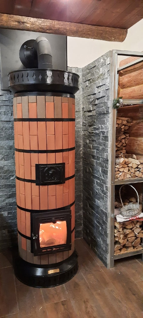 A finished red brick Tundra masonry heater installed against a dark slate heat shield wall, next to a metal firewood storage rack.