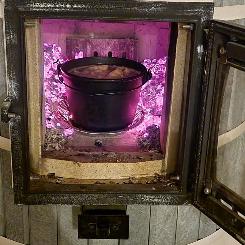 A cast iron dutch oven slow-cooking a stew inside a Siberia masonry heater firebox, surrounded by hot glowing coals.