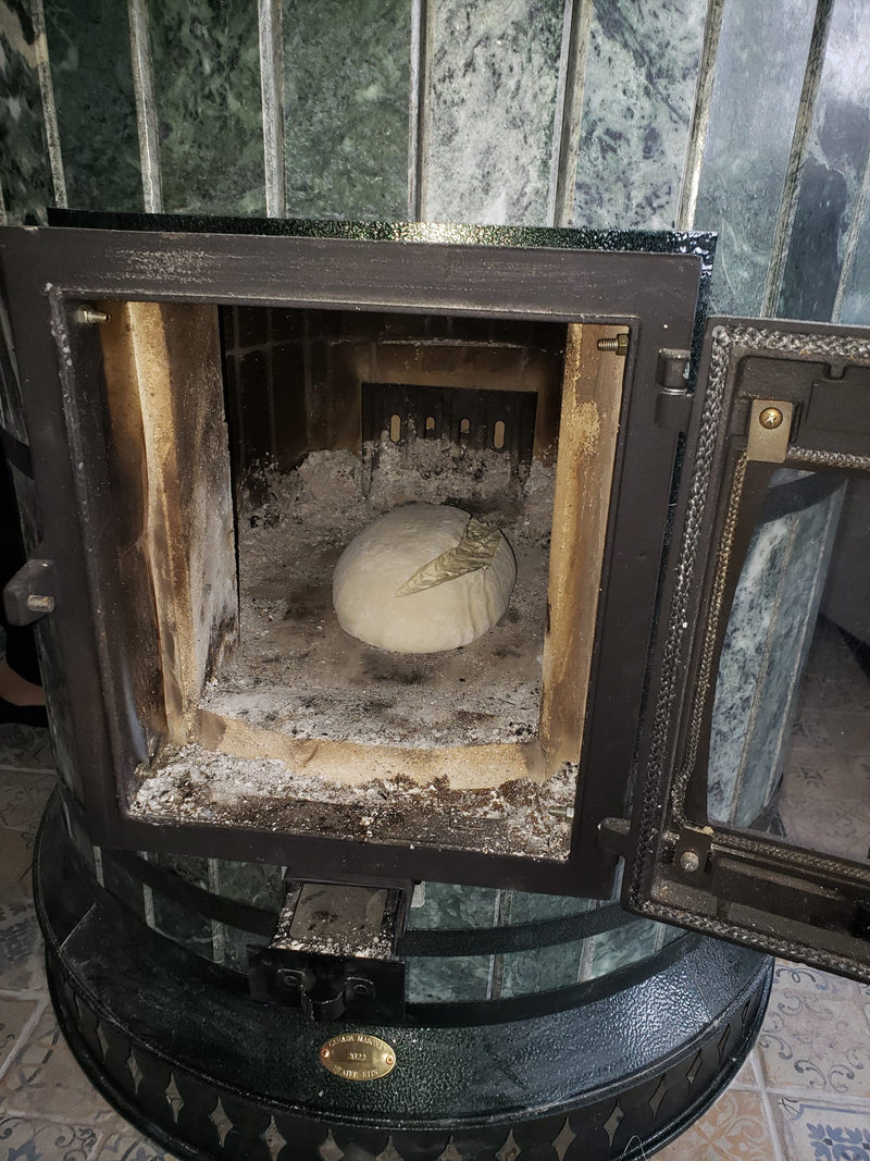 Raw bread dough placed inside the glass firebox door of a green serpentine Siberia masonry heater, utilizing the retained thermal mass for baking.