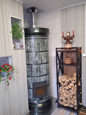 A Siberia green serpentine stone masonry heater installed in a living space next to a black metal firewood storage rack and decorative shelving.