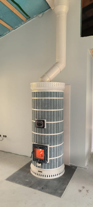 A wood fire burning efficiently inside the glass firebox of a blue Siberia soapstone blue masonry heater kit connected to a white chimney pipe.