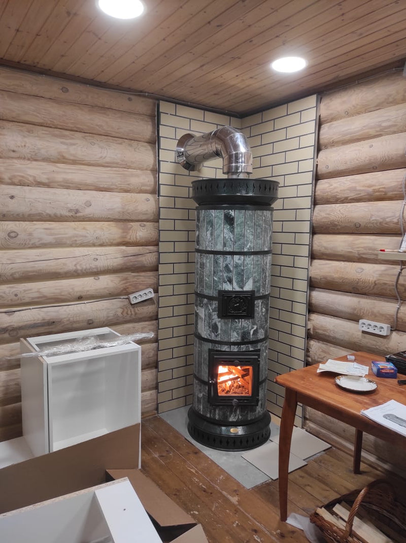A Tundra masonry heater with green serpentine stone cladding installed in a log cabin corner, featuring a beige brick heat shield and stainless steel chimney elbow.