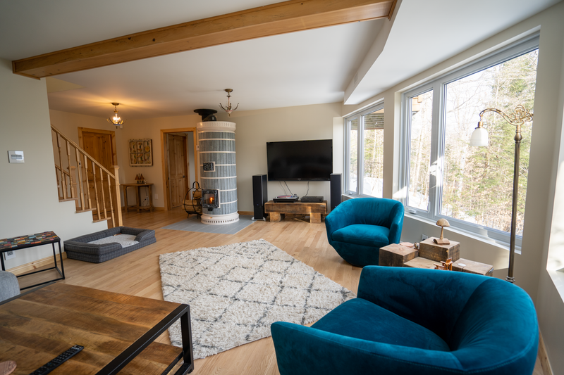 A luxurious living room featuring teal velvet armchairs and large windows, centered around a Siberia soapstone stone masonry heater with white decorative trim.