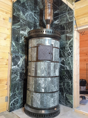 Green masonry saunaheater installed in a corner with matching green stone wall tiling and stainless steel chimney, inside an off-grid cabin
