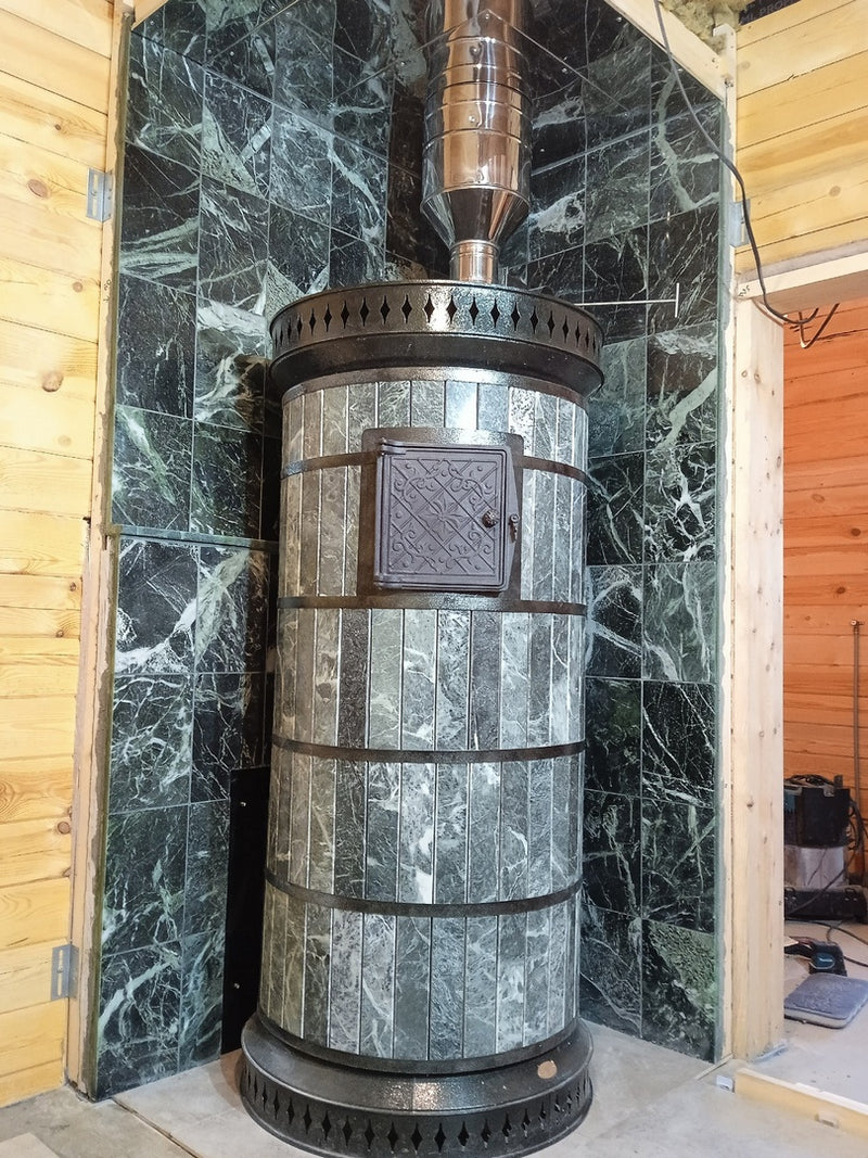 Green masonry saunaheater installed in a corner with matching green stone wall tiling and stainless steel chimney, inside an off-grid cabin
