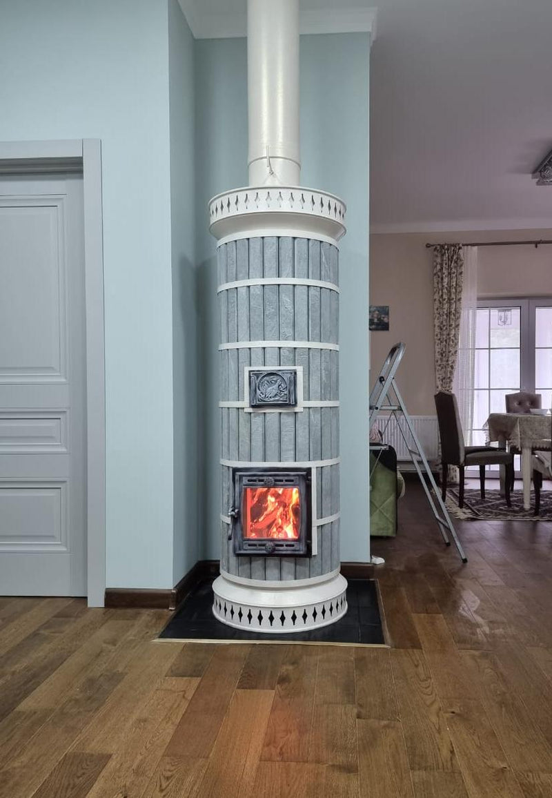 A Tundra masonry heater featuring blue soapstone cladding and contrasting white decorative trim, installed in a bright modern living room next to a white doorway.