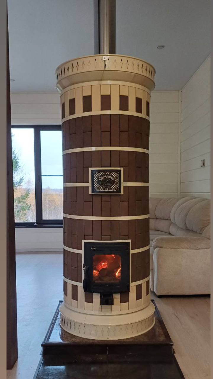 A large DIY Brown Siberia masonry heater serving as the central heating source in a modern living room with white walls and large windows.