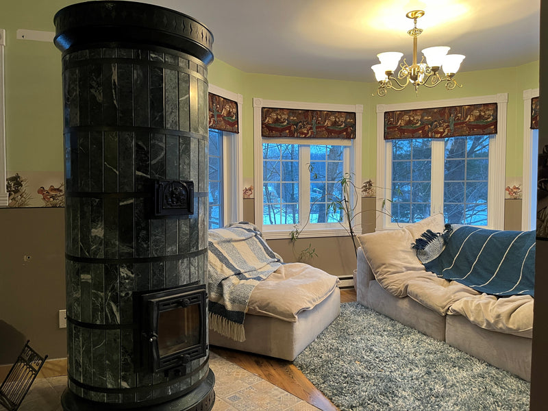 A Siberia green serpentine stone heater serving as the centerpiece of a sunlit living room with bay windows and a blue rug.