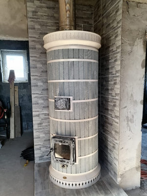 A complete Siberia blue masonry heater kit installed in a tiled corner, showing grey soapstone decorative cladding, cast iron firebox door, and upper cleanout door.