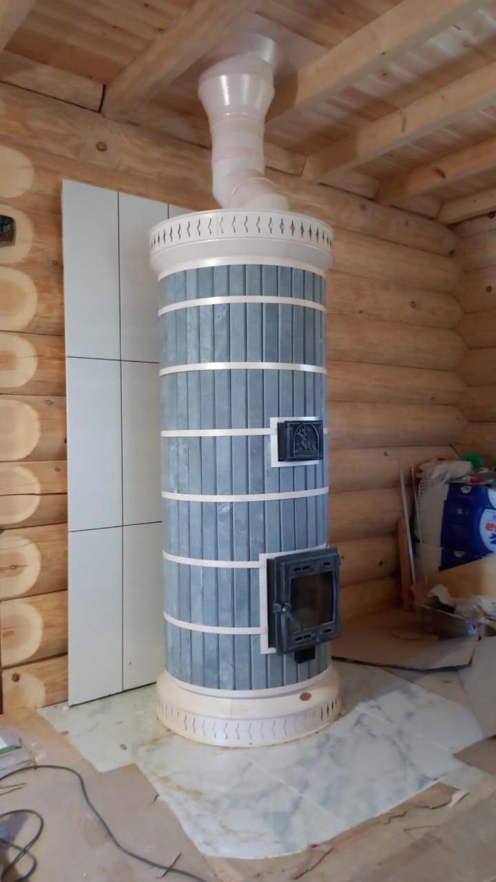 A Siberia soapstone masonry heater kit installed against a rustic timber log wall, showing the side profile and flue connection.