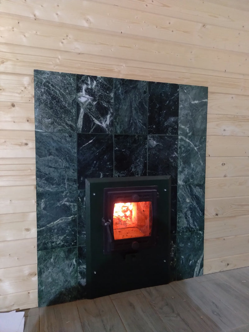 Glass firebox door of green stone sauna heater installed through a wooden wall, surrounded by matching green serpentine tiles