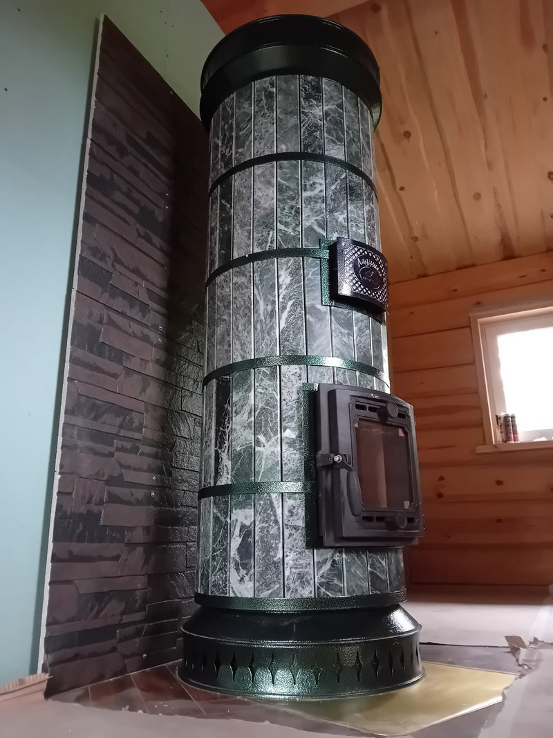 Vertical view of a Tundra green serpentine stone masonry heater installed in a log cabin corner, featuring a dark slate stone heat shield wall.