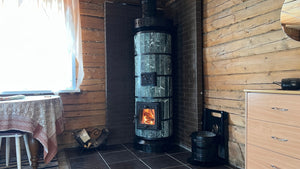 A Tundra green serpentine stone masonry heater installed in a tight corner with a dark brick heat shield, positioned next to a wooden dresser.