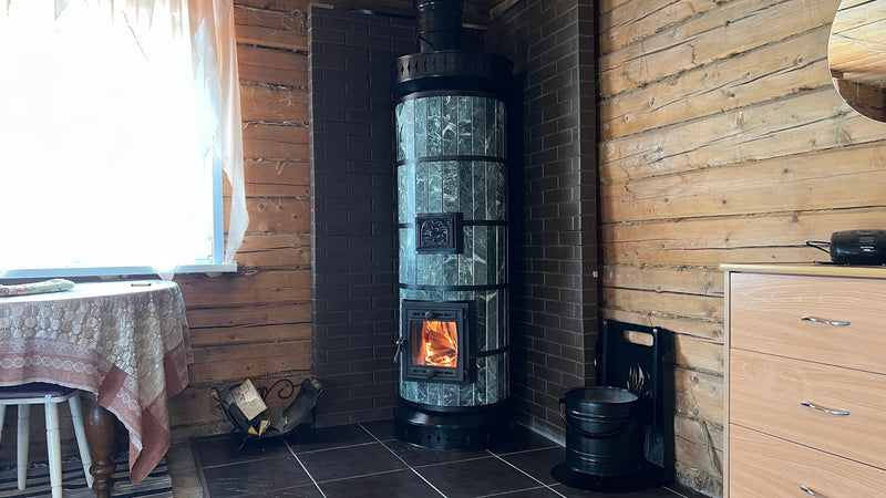A Tundra green serpentine stone masonry heater installed in a tight corner with a dark brick heat shield, positioned next to a wooden dresser.