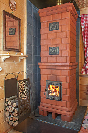 Finished Volga masonry heater installed in a wooden log cabin, featuring decorative relief tiles above the firebox and a metal log holder.