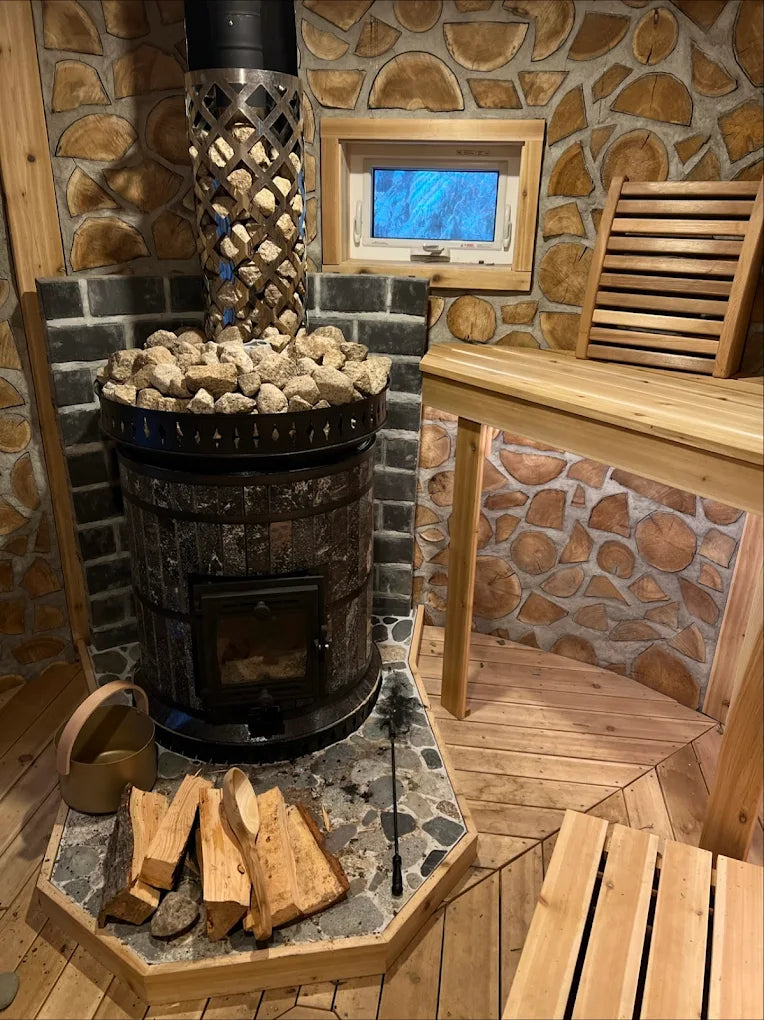 Full view of a black stone Volcano masonry wood-fired sauna stove featuring a stone-filled mesh chimney basket for maximum heat retention in a rustic sauna.